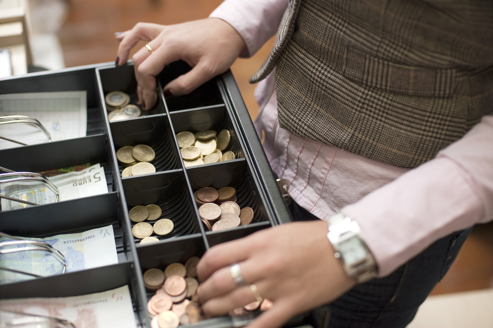 Imagem mostra as mãos de uma mulher mexendo em moedas na gaveta aberta de um caixa de loja