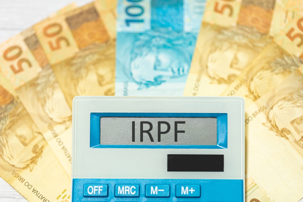 Imagem mostra calculadora com a sigla IRPF escrita na tela e, ao fundo, notas de 50 reais e uma nota de 100 reais dispostas como um leque