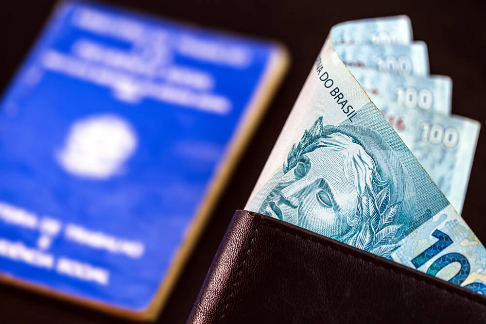 Imagem mostra uma carteiro preta de couro com notas de 100 reais dobradas ao meio e, ao fundo, uma carteira de trabalho