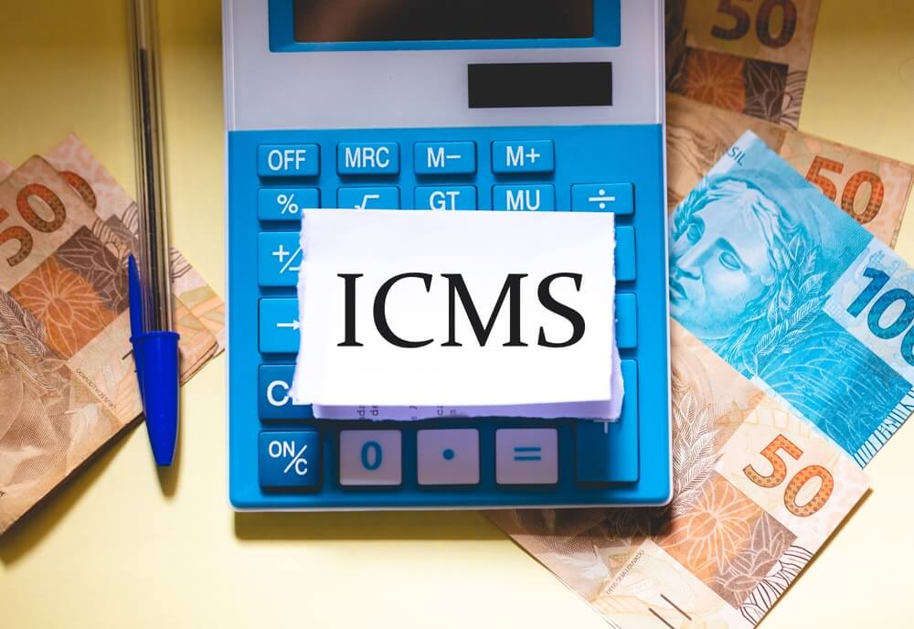 Imagem mostra caneta, notas de dinheiro e uma calculadora com um papel escrito ICMS em cima.