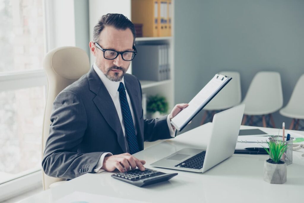 Um homem em seu escritório, com um computador na mesa, na mesma mesa uma calculadora onde ele está mexendo e em sua outra mão uma prancheta