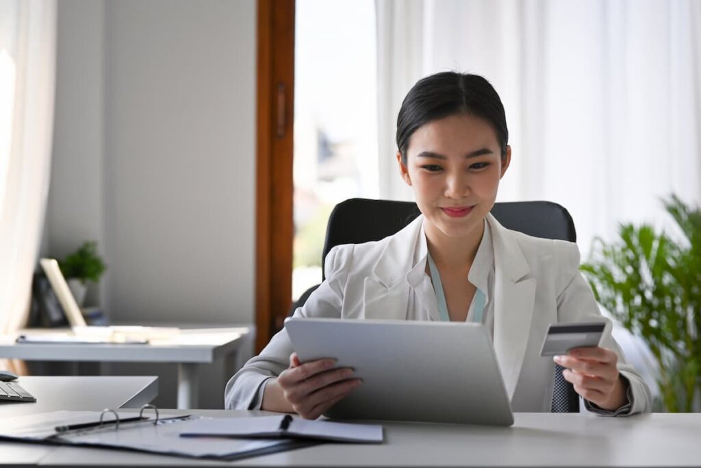 Uma mulher de blazer branco, em uma mesa de escritório, com um tablet em sua mão e um cartão em outra