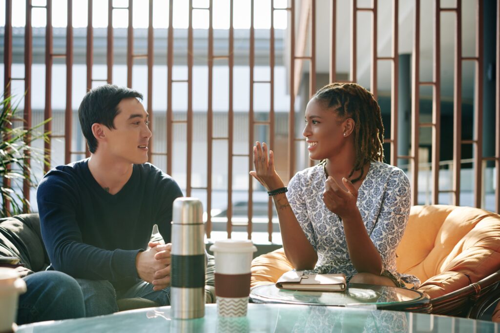 Homem e mulher conversam em espaço com poltronas e um mesa com um copo de café e uma garrafa térmica