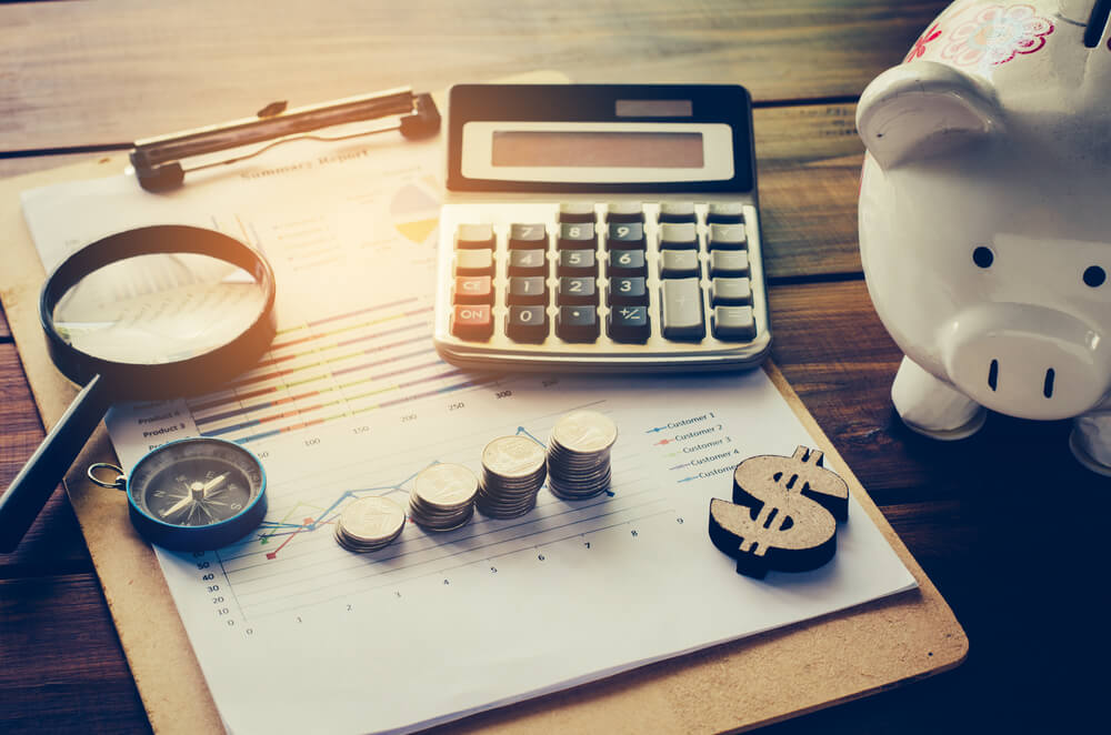 Imagem mostra elementos que remetem a uma empresa de serviços financeiros, como um cofre de porquinho, uma calculadora, moedas, uma lupa, uma bússola e um cifrão de madeira
