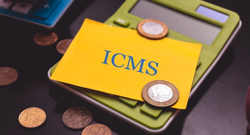 Imagem mostra calculadora com um papel em cima escrito ICMS e moedas em volta