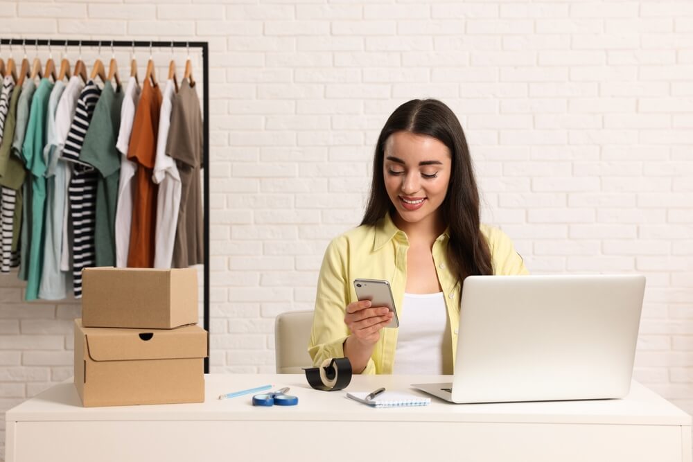 Empreendedora que vende roupas em frente a um laptop enquanto mexe no celular, com uma arara com camisetas ao fundo e caixas ao lado dela na mesa