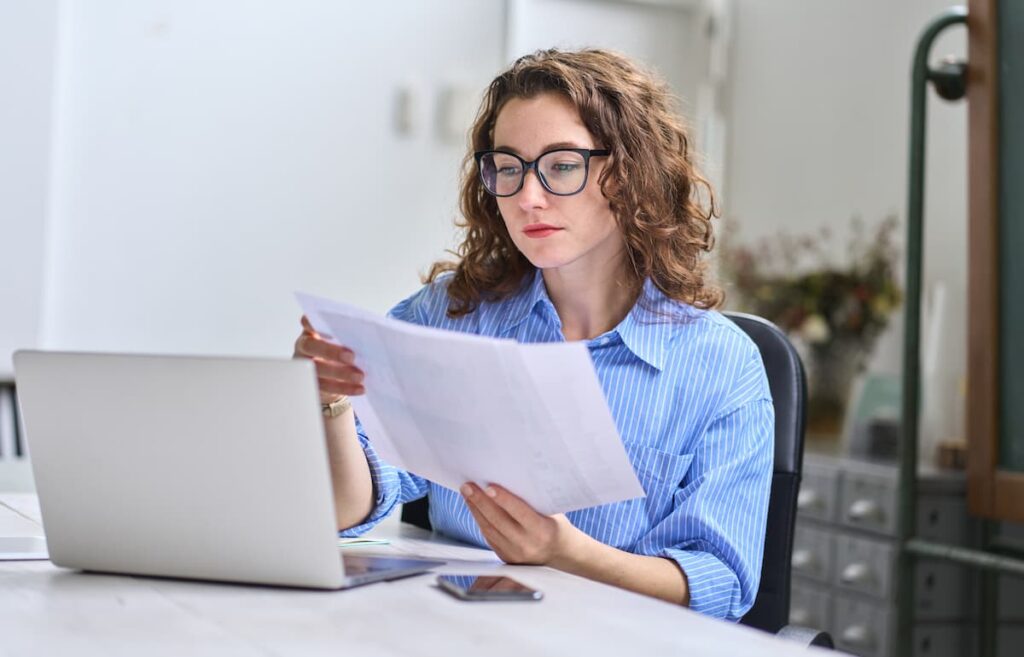 Um mulher com roupa social azul e óculos preto, segurando em suas mãos um papel e com seu computador na sua frente