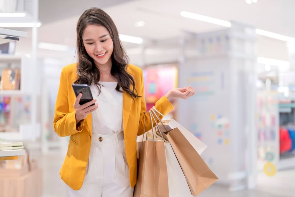 Uma mulher de blazer amarelo usa o telefone enquanto carrega sacolas de compras em uma loja

