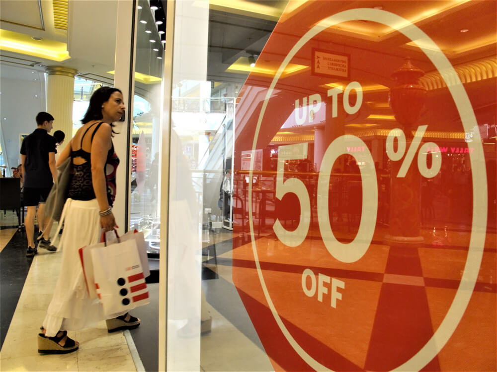 Consumidores passando por uma loja anunciando uma promoção com até 50% de desconto
