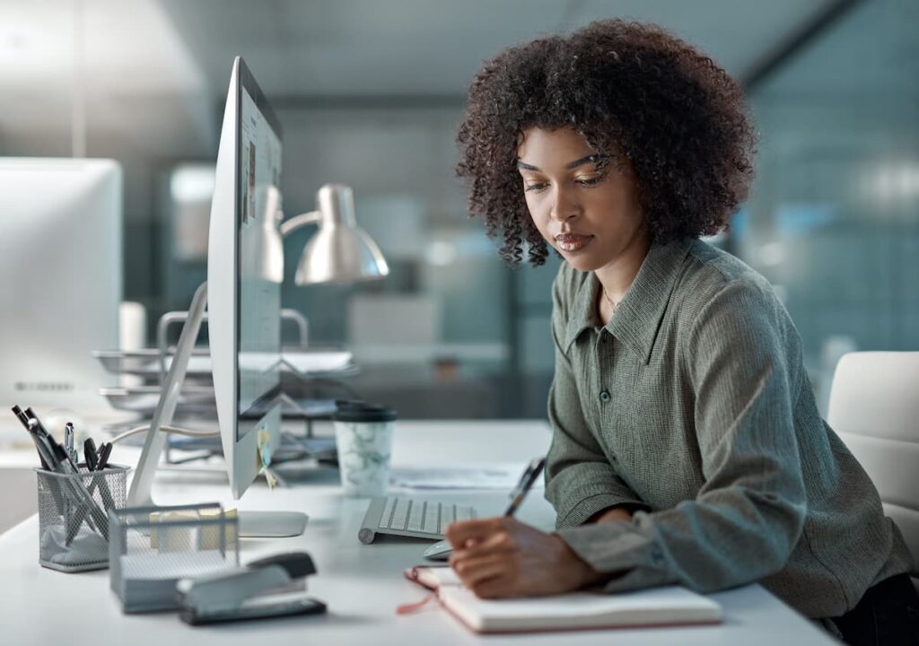 Uma mulher no escritório, passando informações vistas em seu computador para o caderno